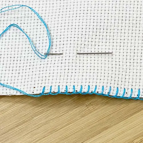 Edge of white aida cloth sewn with blanket stitch using blue embroidery floss all resting on a bamboo surface