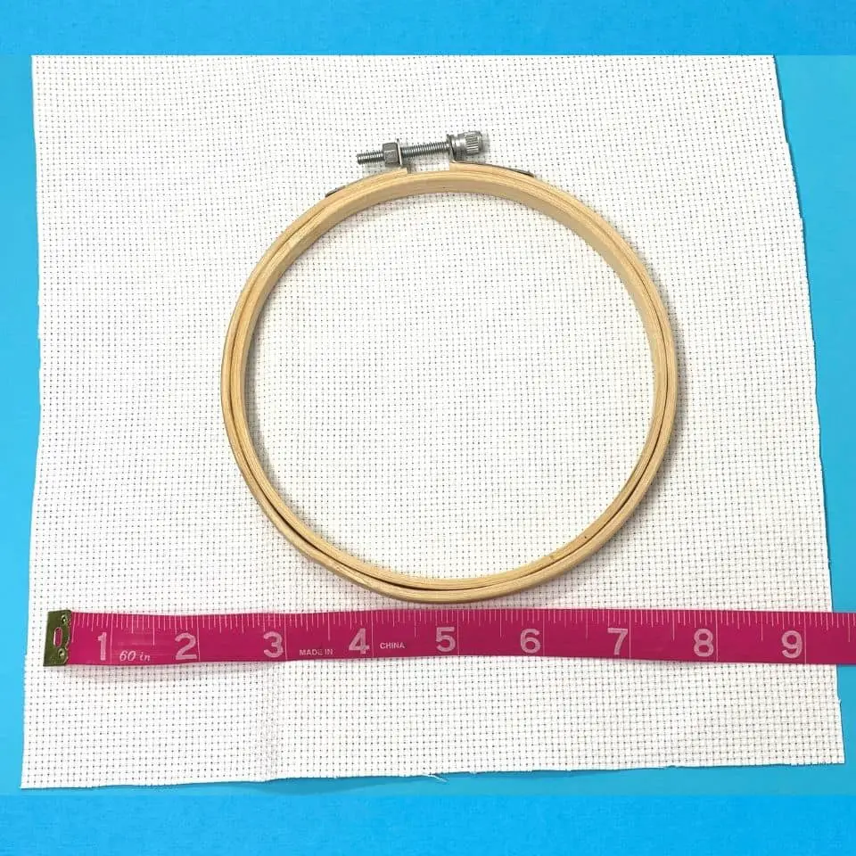 Bamboo embroidery hoop on top of white aida cloth with a pink measuring tape showing the aida cloth is nine and a half inches all over a light blue background 