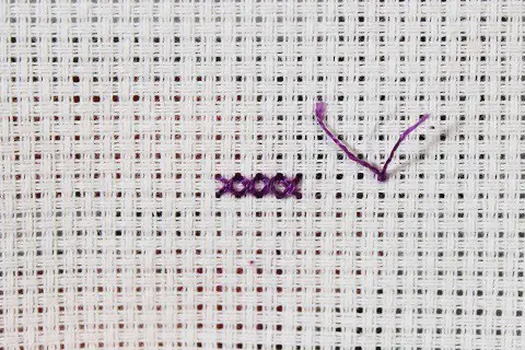 4 Cross stitches in purple thread on white aida cloth with an away knot positioned 4 fabric squares to the right of the stitches