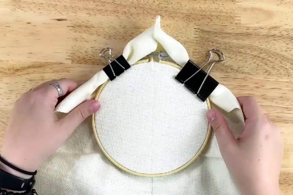An embroidery hoop with fabric has binder clips at the top-right and top-left holding extra, rolled up fabric while two hand hold the hoop on a flat wood surface
