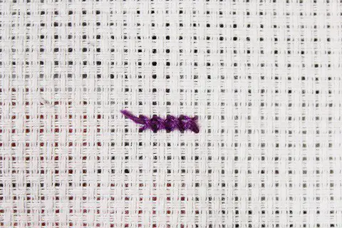 a short row of cross stitches with the tail end tucked under the front of the stitches done in purple embroidery floss on white aida