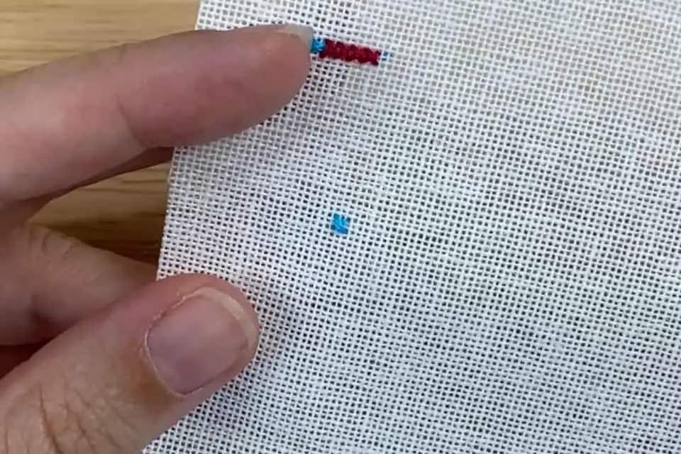 A single blue cross stitch is on a small piece of white linen, held in a left hand