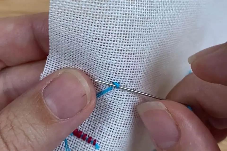 A single blue cross stitch, viewed from the back, on white aida cloth. A needle is passing under the stitch, showing fingers holding the needle and fabric