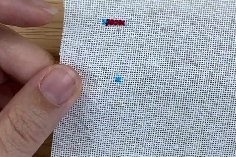 A single cross stitch viewed from the front in blue embroidery floss on white aida. A left thumb and forefinger are holding the fabric.