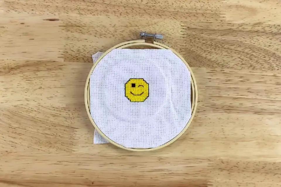 A wink emoji cross stitch project is placed inside a bamboo embroidery hoop, leaving the edges of the fabric showing inside the hoop, on a flat wood background.