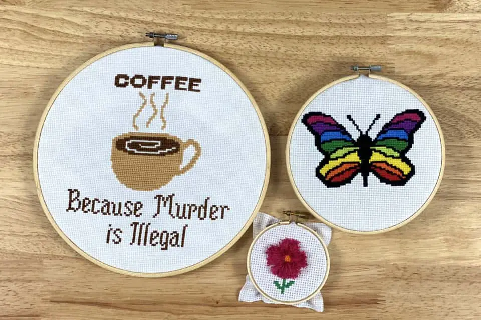 3 cross stitch embroidery projects, the first in a large, wood  embroidery hoop with a cup off coffee in the center that reads coffee, because murder is illegal, the second in a medium, wooden, embroidery hoop with a butterfly that has rainbow wings, the third in a small, wood embroidery hoop with a pink flower stitched in the center