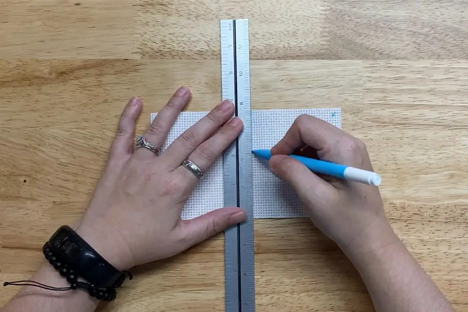 left hand holding a ruler over a small piece of aida cloth with right hand writing on the cloth with a water erasable pen