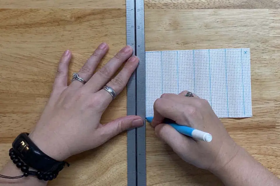 left hand holds a ruler over aida fabric, right hand writes with a water soluble marker