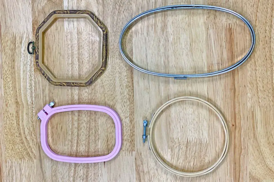 In the top left is a hexagon embroidery hoop, to the top right is a metal, oval embroidery hoop, to the bottom left is a plastic, square embroidery hoop and in the bottom right is a circular, wood embroidery hoop, all laying on a flat, wood background