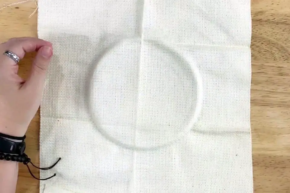 Embroidery fabric with an impression left from an embroidery hoop, placed on a flat wood surface. A left hand is resting on the surface to the left of the fabric.