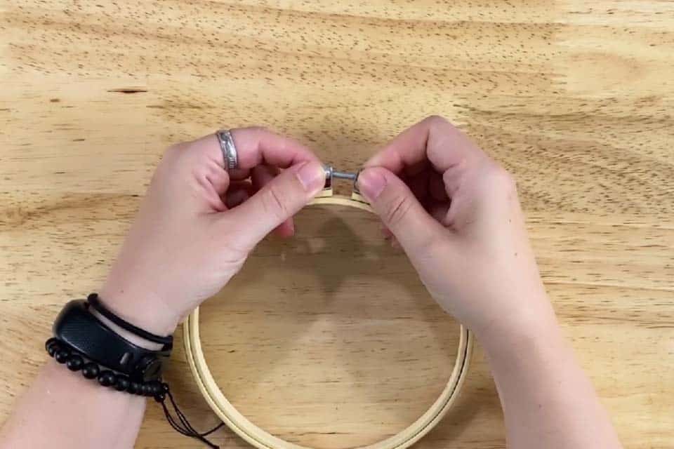 Two hands loosening the screw on a bamboo embroidery hoop over a flat wood background.