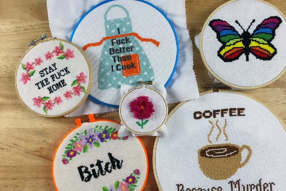 Pile of various finished cross stitch projects in multiple sizes of embroidery hoops against a flat, wood background