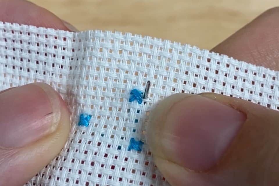 A needle is poking through a small piece of white aida cloth with 3 blue cross stitches on it, while held by a couple of fingers. The background is blurred out.