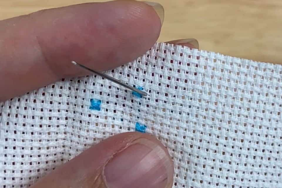 A needle is getting pushed from the back through a small piece of white aida cloth with three blue cross stitches on it, while held in a left hand
