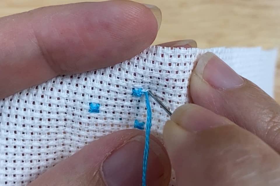 A needle is getting pushed through from front to back of a small piece of white aida fabric with 3 blue cross stitches on it as a few fingers are visible of a left hand, holding the fabric