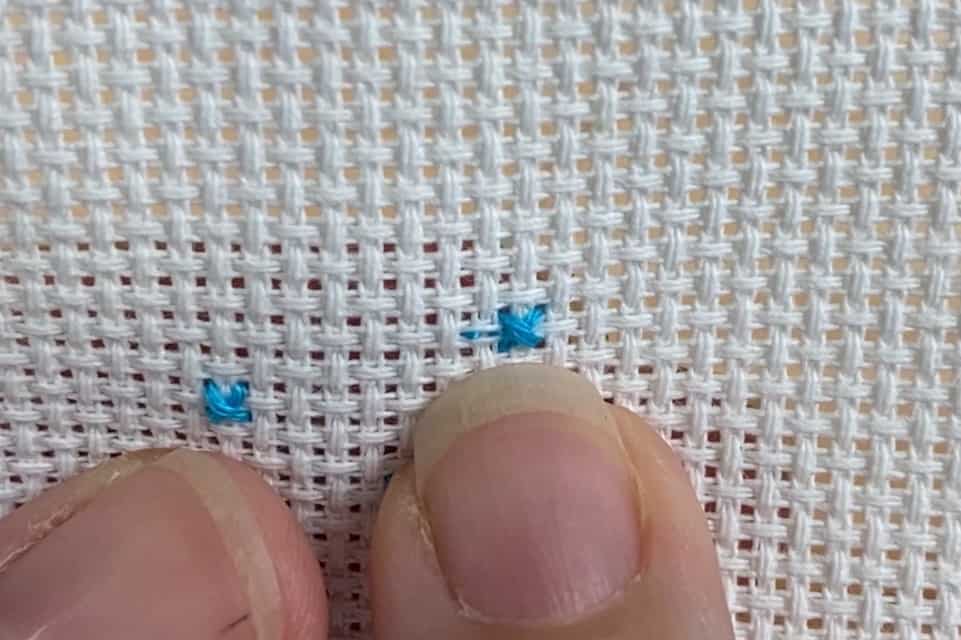 A finger is pointing to a small blue cross stitch on white aida cloth