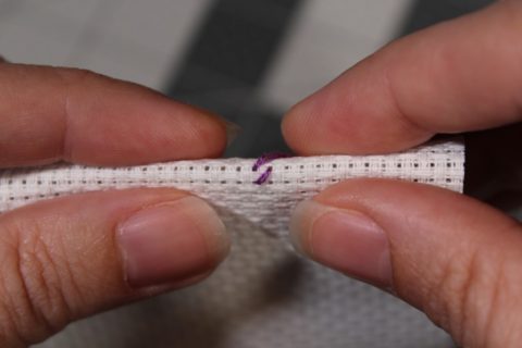 two hands pinching folding aid cloth showing a row of half cross stitches in purple embroidery on white aida