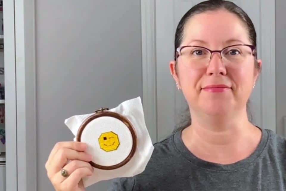 A 3 inch cross stitch of a wink emoji is framed in a faux wood embroidery hoop, held next to Sarah's smiling face.