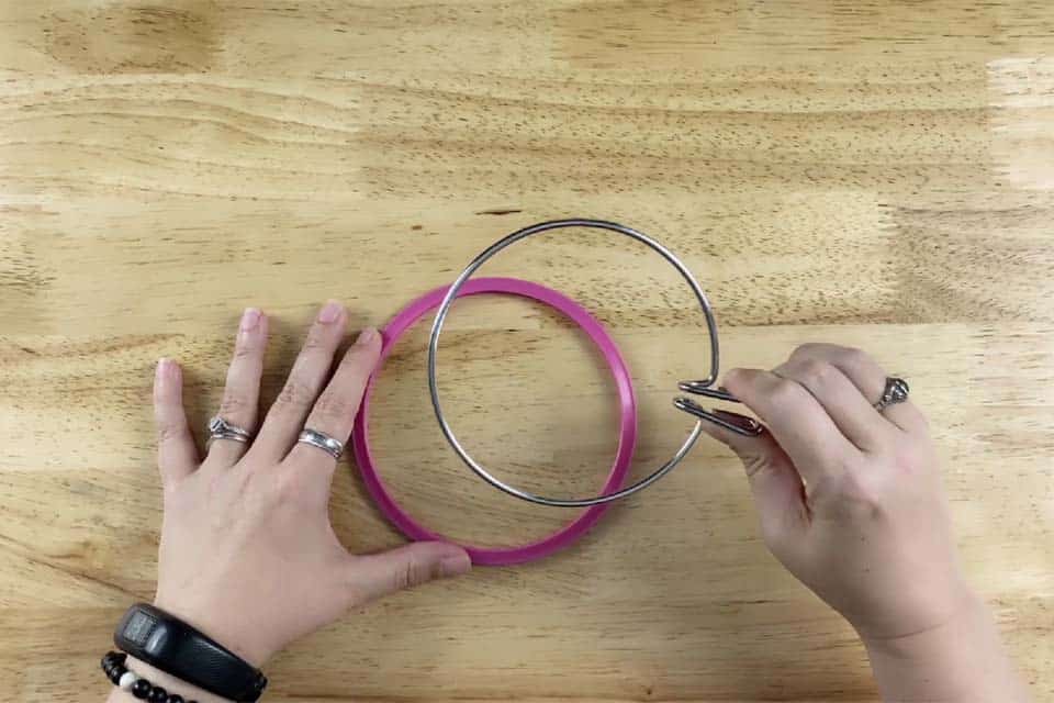 A spring tension embroidery hoop with the inside metal hoop held in a right hand squeezing the metal handles and the pink, plastic outer hoop resting on a flat wood surface with a left hand touching it