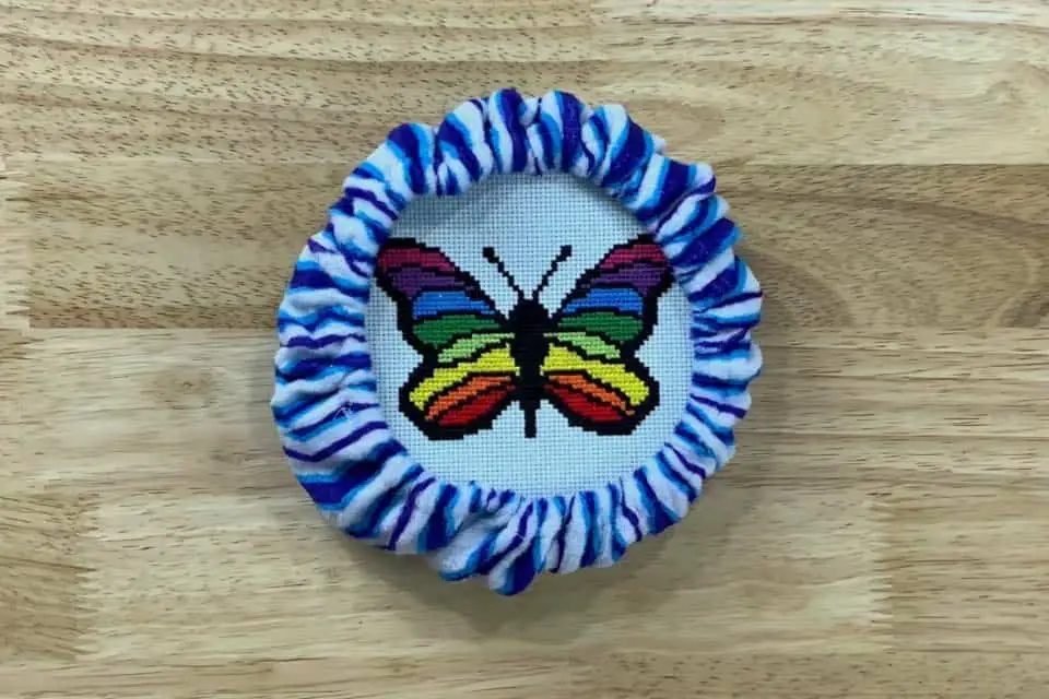 Blue and white striped steering wheel cover is covering an embroidery hoop with a rainbow butterfly cross stitch inside