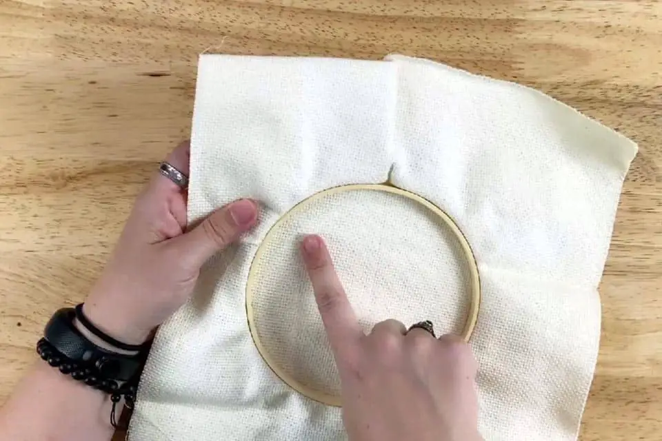 Embroidery hoop with fabric is held upside down in left hand, with right index finger pointing at the inside of the emrboidery hoop. A plain wood surface is in the background.