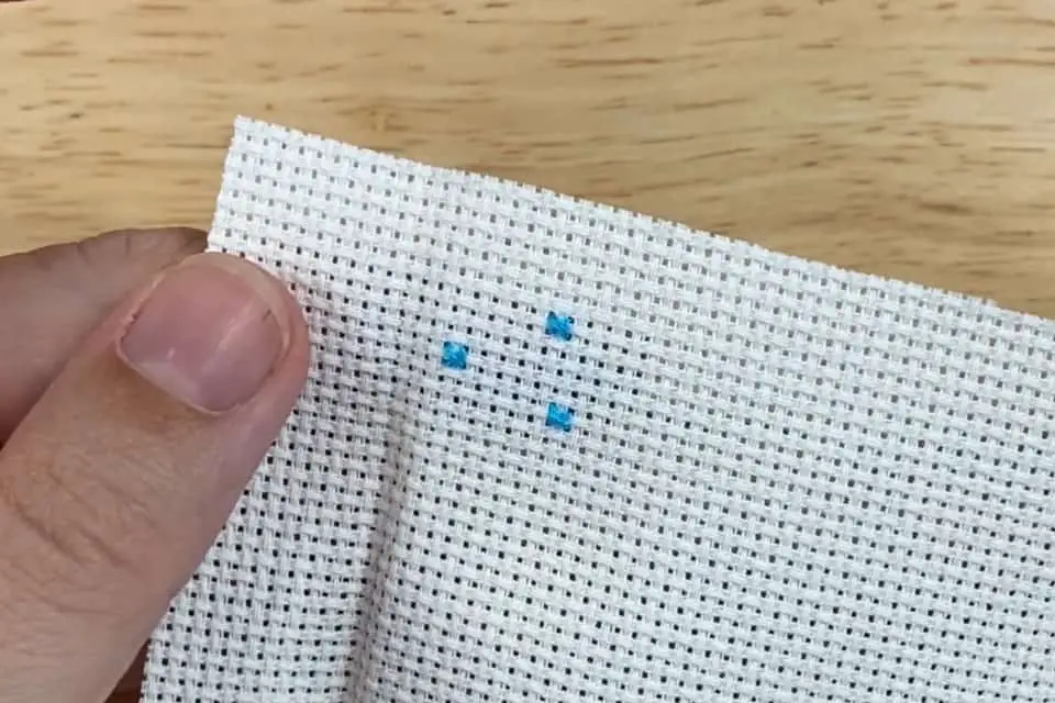 Three single, blue cross stitches one a small piece of white, aida cloth, held by a left thumb and forefinger over a wood background