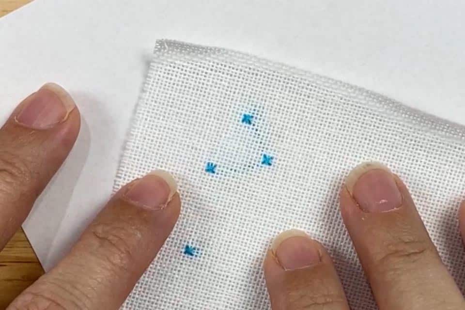 A white piece of linen with 4 small blue cross stitches where the floss connecting the stitches is visible through to the front of the fabric. The linen is held down by 4 fingers.