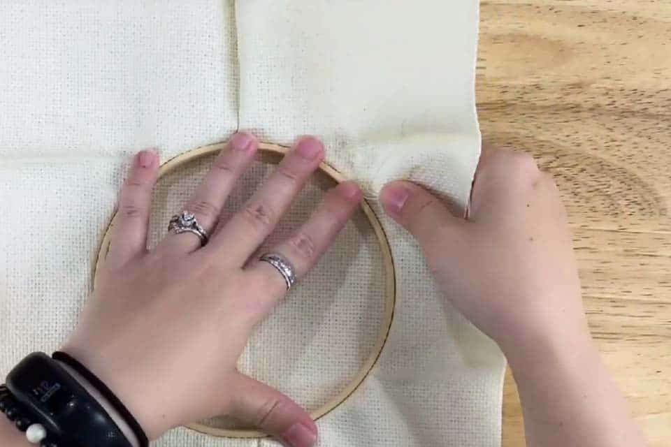 An embroidery hoop with fabric is held upside down on a flat wood surface with a left hand while the right hand tugs at the edge of the fabric