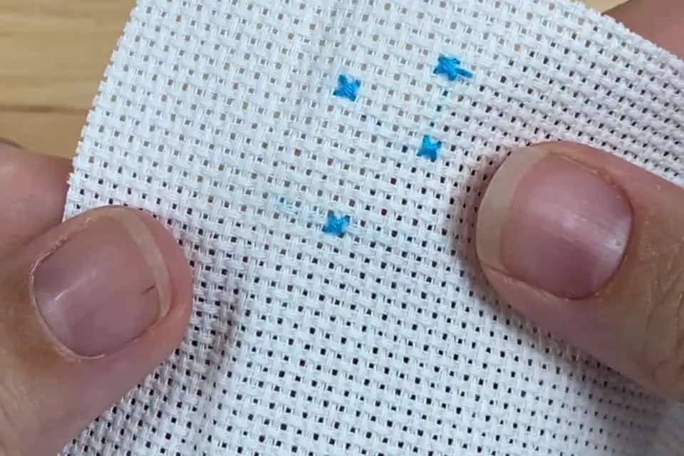 Four small blue cross stitches on a small piece of white aida cloth. Two thumbs are visible, holding the cloth.