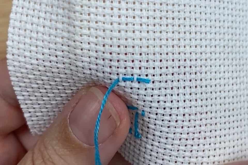 Blue embroidery floss is woven through two fabric squares on white aida cloth. A left thumb is visible holding the fabric.