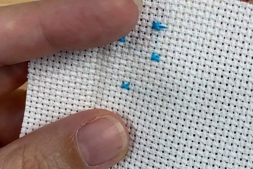 Three small blue cross stitches are on a small piece of white aida fabric held by a left hand