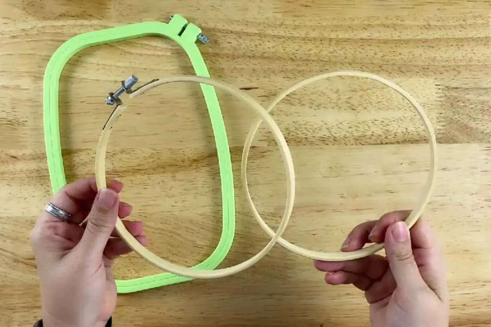 A wood embroidery hoop separated with the outer hoop held in a left hand and the inner hoop held in a right hand with a green, square plastic embroidery hoop in the background on top of a flat, wood surface