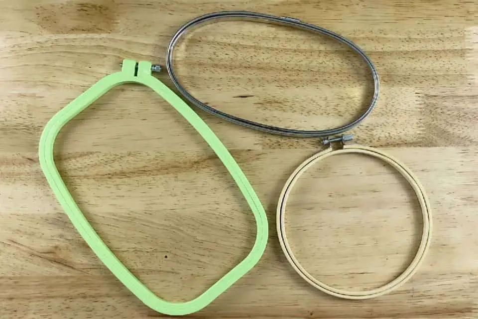 Three embroidery hoops, a metal hoop at the top, and green, plastic hoop to the left and a wood hoop to the right, all on top of a flat wood surface