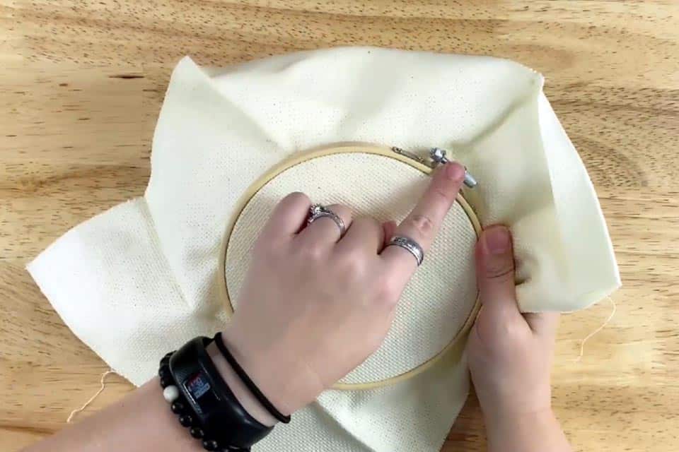 Embroidery hoop with fabric held in right hand as the hoop hardware is in a 1 o'clock position. Index finger on left hand points to hardware. Wood surface in background.