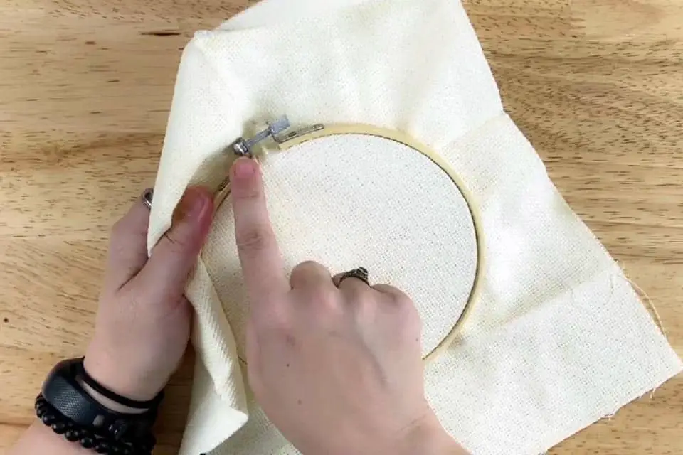 Embroidery hoop with fabric held in left hand as the hoop hardware is in a 10 o'clock position. Index finger on right hand points to hardware. Wood surface in background.